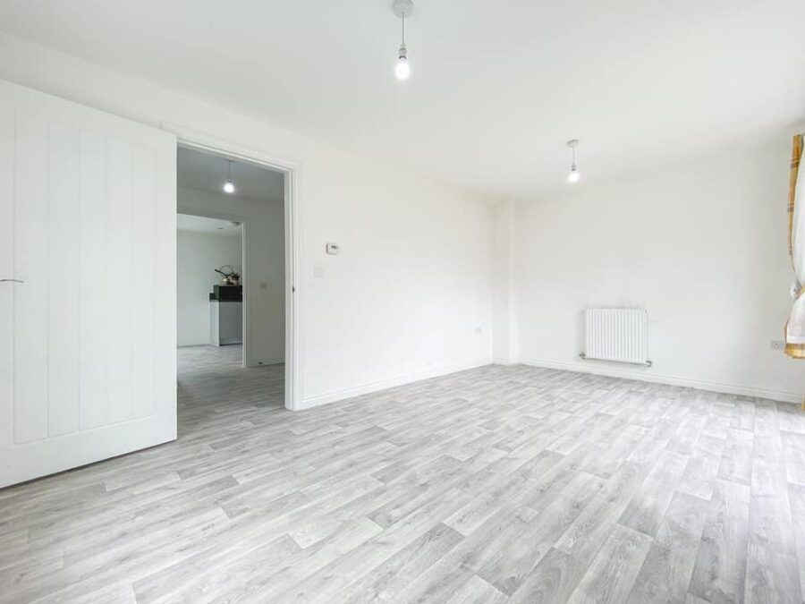 A wide-angle view of a bright, empty room with white walls and light grey wood-effect vinyl plank flooring. An open white door leads to another room, and two simple pendant lights hang from the ceiling.