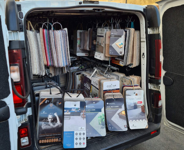 The open back of a white professional van filled with rows of hanging carpet and flooring samples, including brands like Cormar and Lifestyle Floors, parked in a residential area.