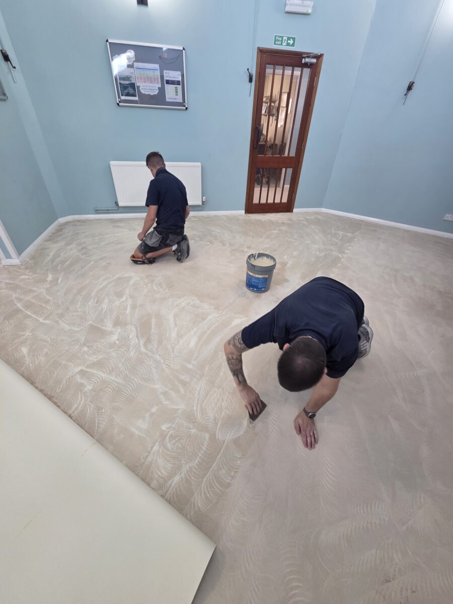 Two Avana Flooring fitters in blue uniforms kneeling on a subfloor, expertly applying adhesive with spreaders to prepare for a sheet vinyl installation.