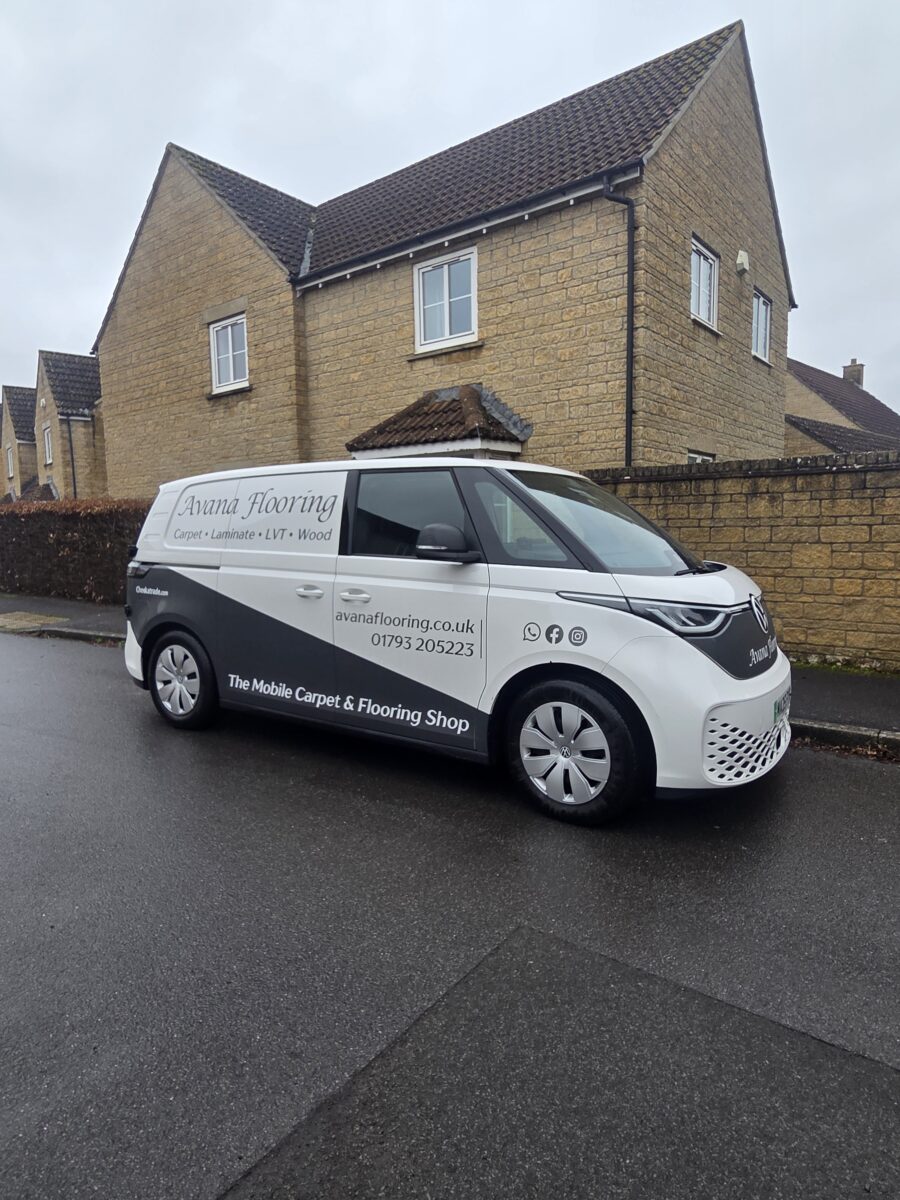 A branded white and black Volkswagen ID. Buzz electric van for Avana Flooring, "The Mobile Carpet & Flooring Shop," parked on a the side of the road in front of a modern yellow-brick house in Calne.