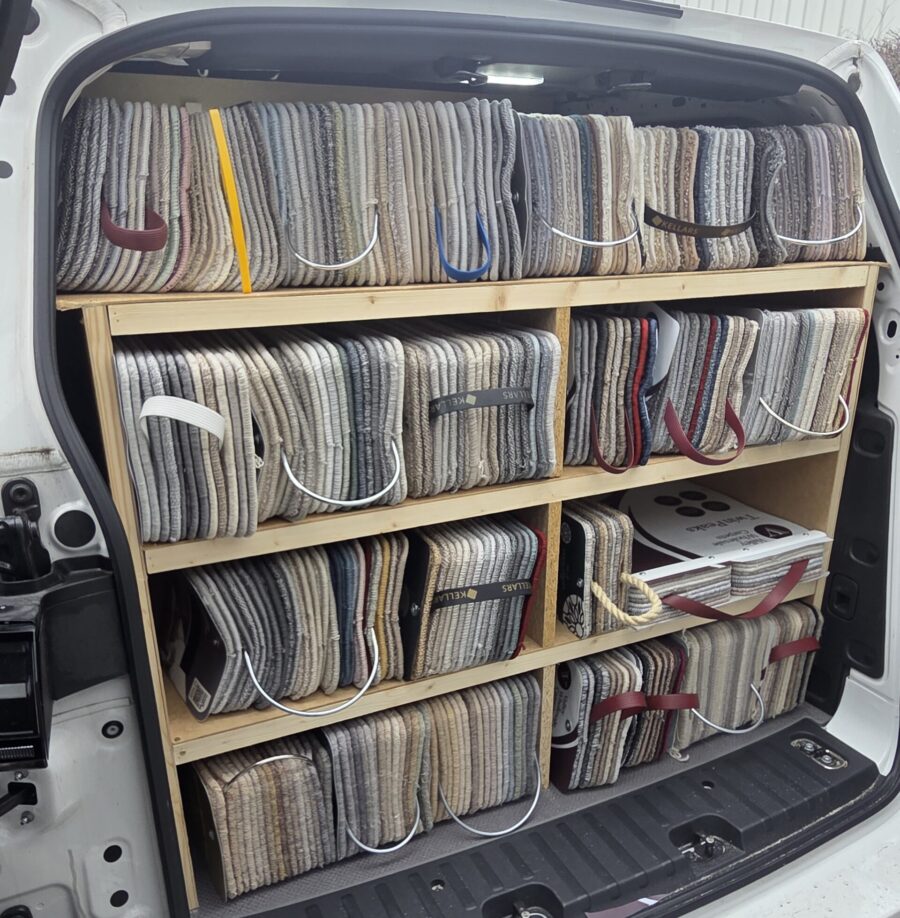 Interior of an Avana Flooring van featuring organised wooden shelving units filled with a wide variety of carpet sample books and swatches for home consultations.