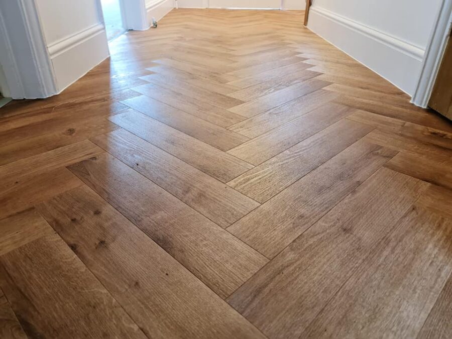 A close-up view of a professional Karndean oak-effect herringbone LVT floor installation in a hallway, showing the detailed wood grain texture and precise fitting against white skirting boards.