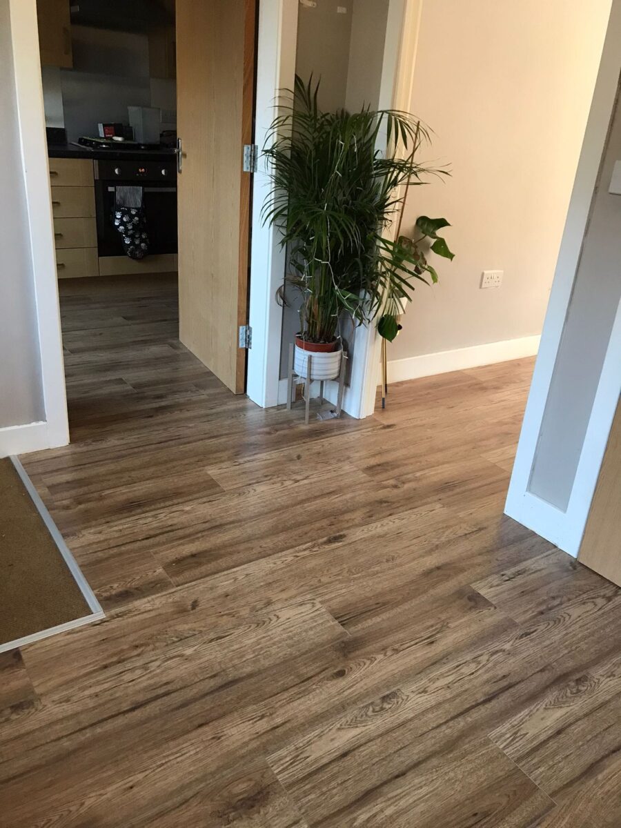A wide-angle view of a residential hallway featuring a professional installation of mid-tone oak-effect laminate flooring. The floor flows seamlessly into an open kitchen area, complemented by white skirting boards and a potted plant.