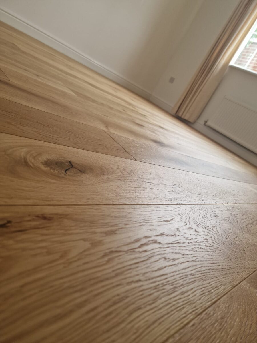 A low-angle, close-up photograph of natural oak engineered wood flooring planks, showing detailed wood grain, knots, and a textured matte finish in a bright room.