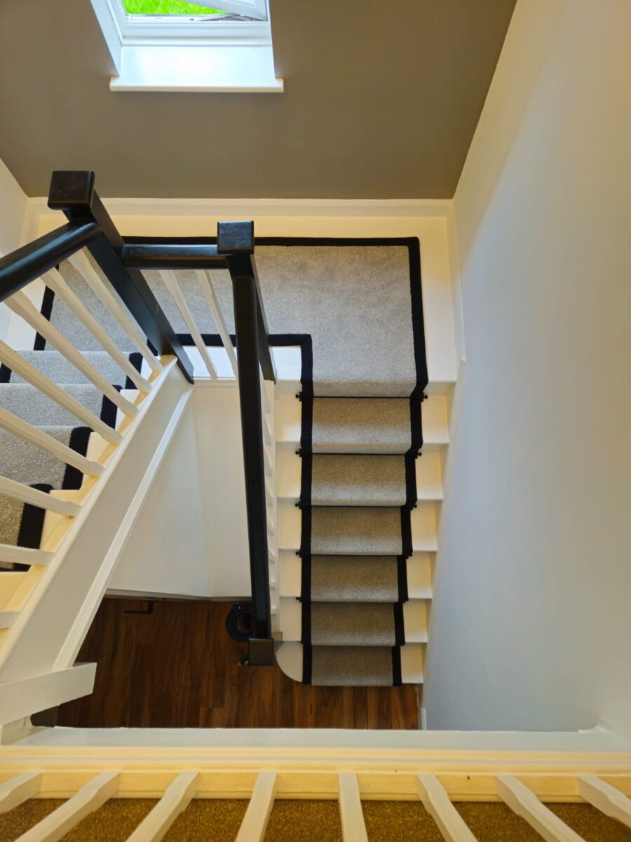 A high-angle view looking down a white staircase with a light stone carpet runner featuring a sharp black border, matching the black handrails and dark wood flooring below.