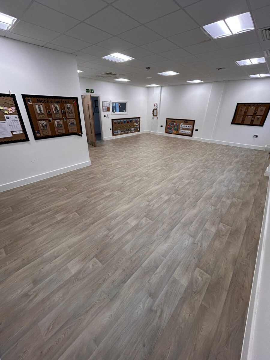 A wide-angle view of a large, bright commercial room featuring a professional installation of wood-effect sheet vinyl flooring with a seamless finish.