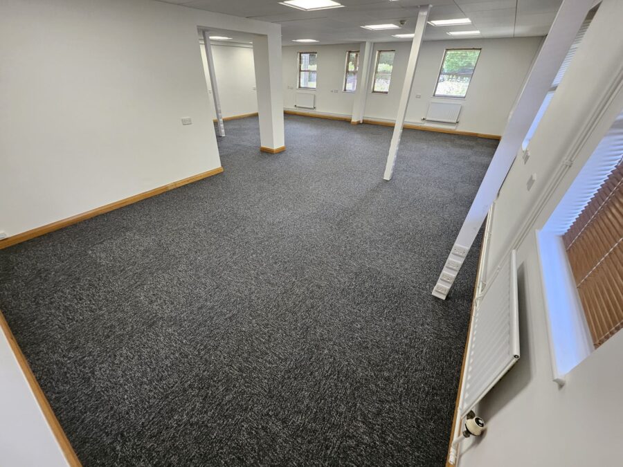 A high-angle, wide-view of a spacious, white-walled office featuring a professional installation of grey-speckled carpet tiles with white structural pillars and oak-effect skirting.