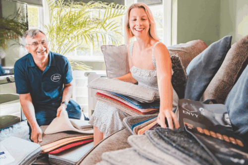 A friendly Avana Flooring expert in a branded polo shirt assisting a smiling customer in her sunlit living room as she browses through a variety of plush carpet samples.